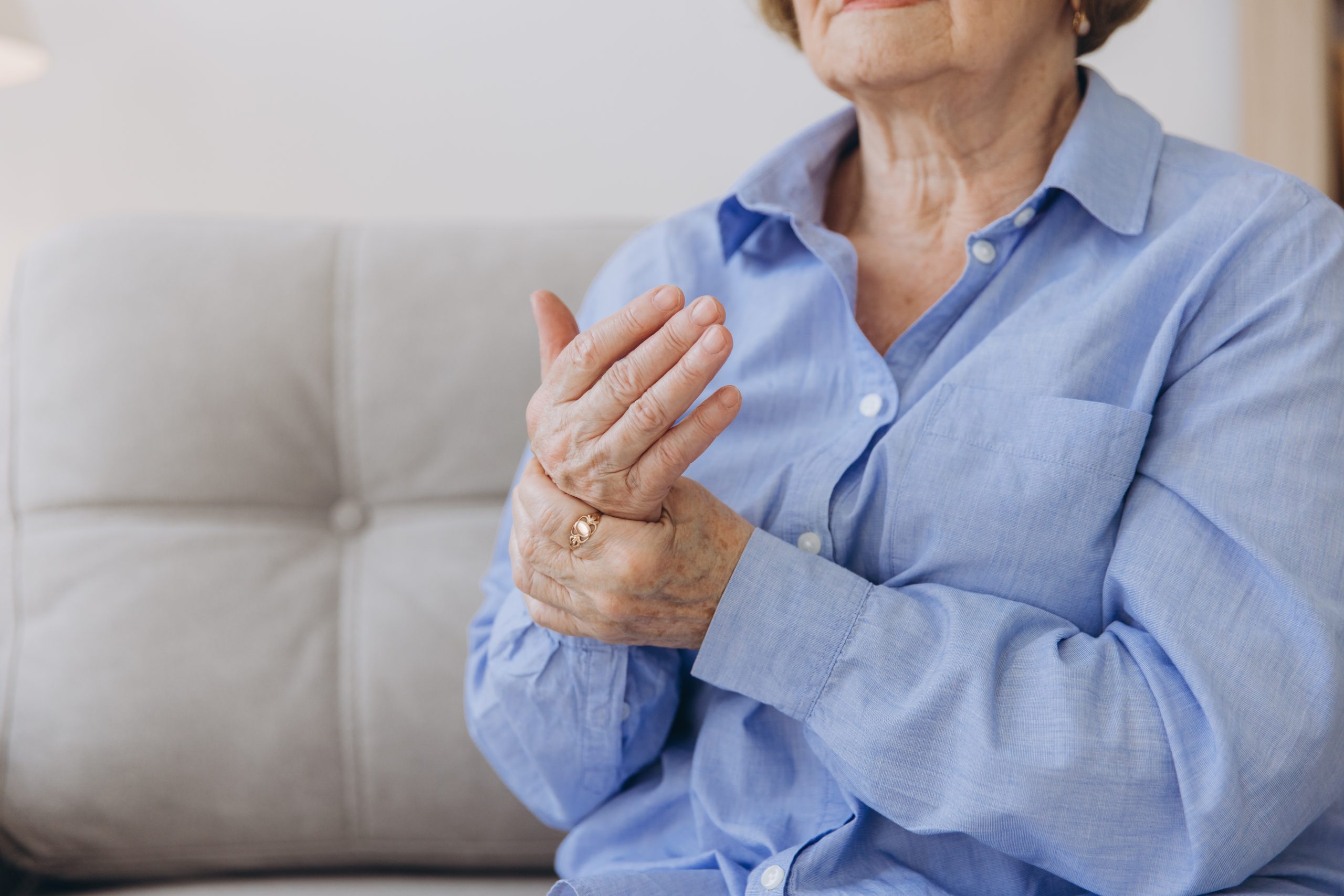 An elderly person wearing a blue shirt is holding their hand, possibly massaging it, while sitting on a light-colored sofa.