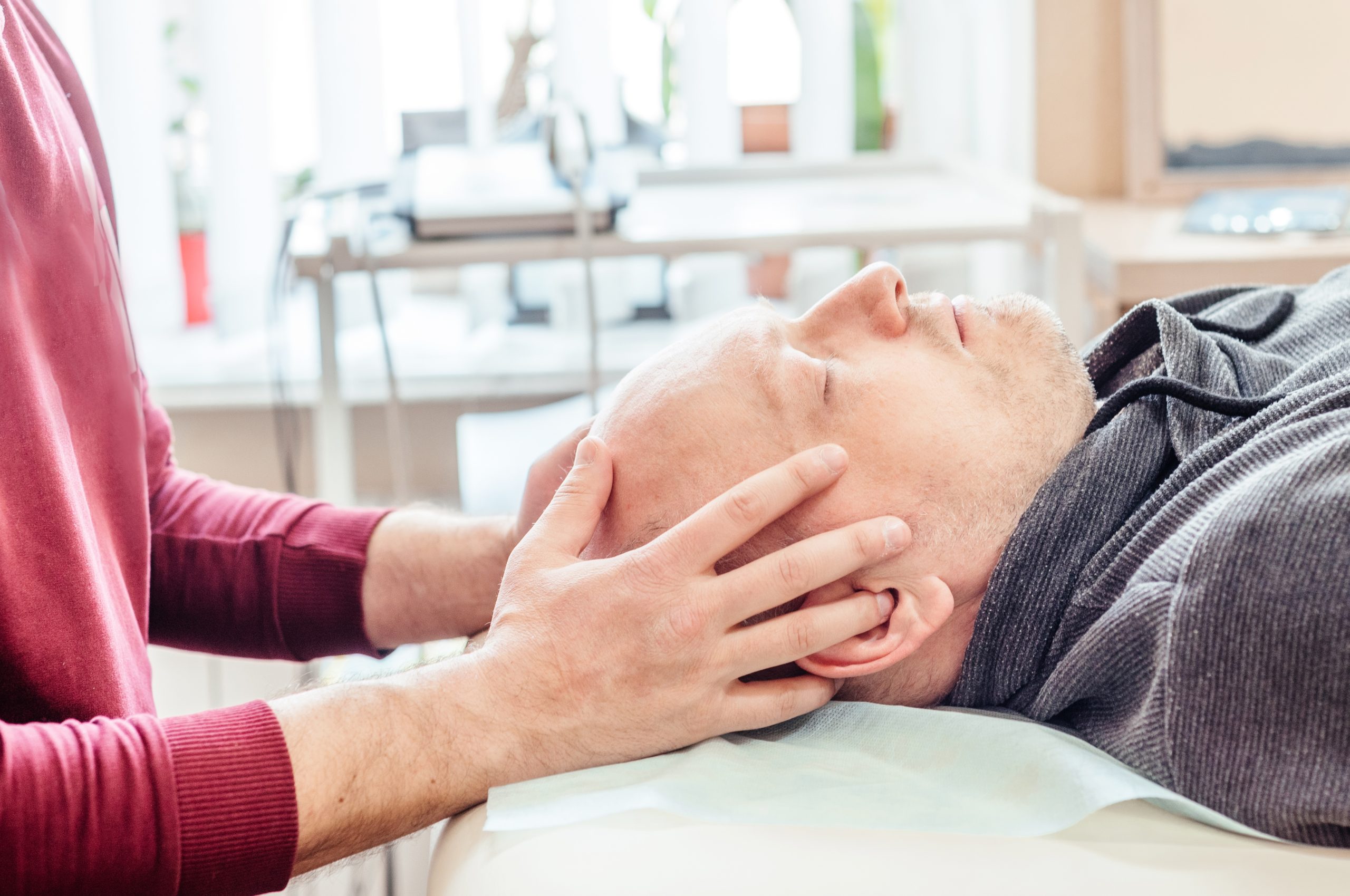 Person receiving a head massage, lying relaxed on a cushioned surface. Hands gently cradle the head. Brightly lit room with medical or spa equipment in the background.