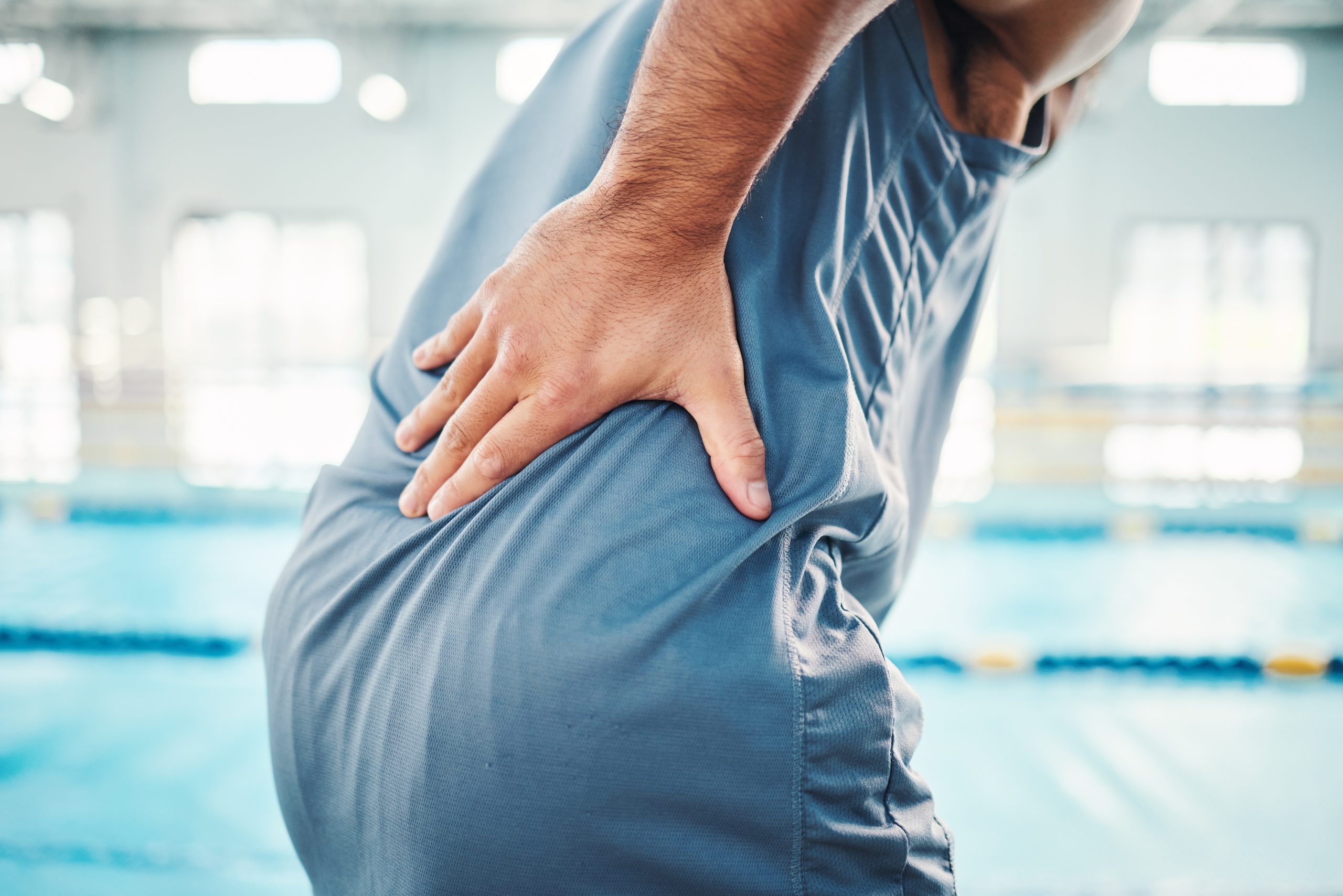 A person's hand rests on their lower back, suggesting discomfort, while standing in a bright, indoor swimming pool setting.