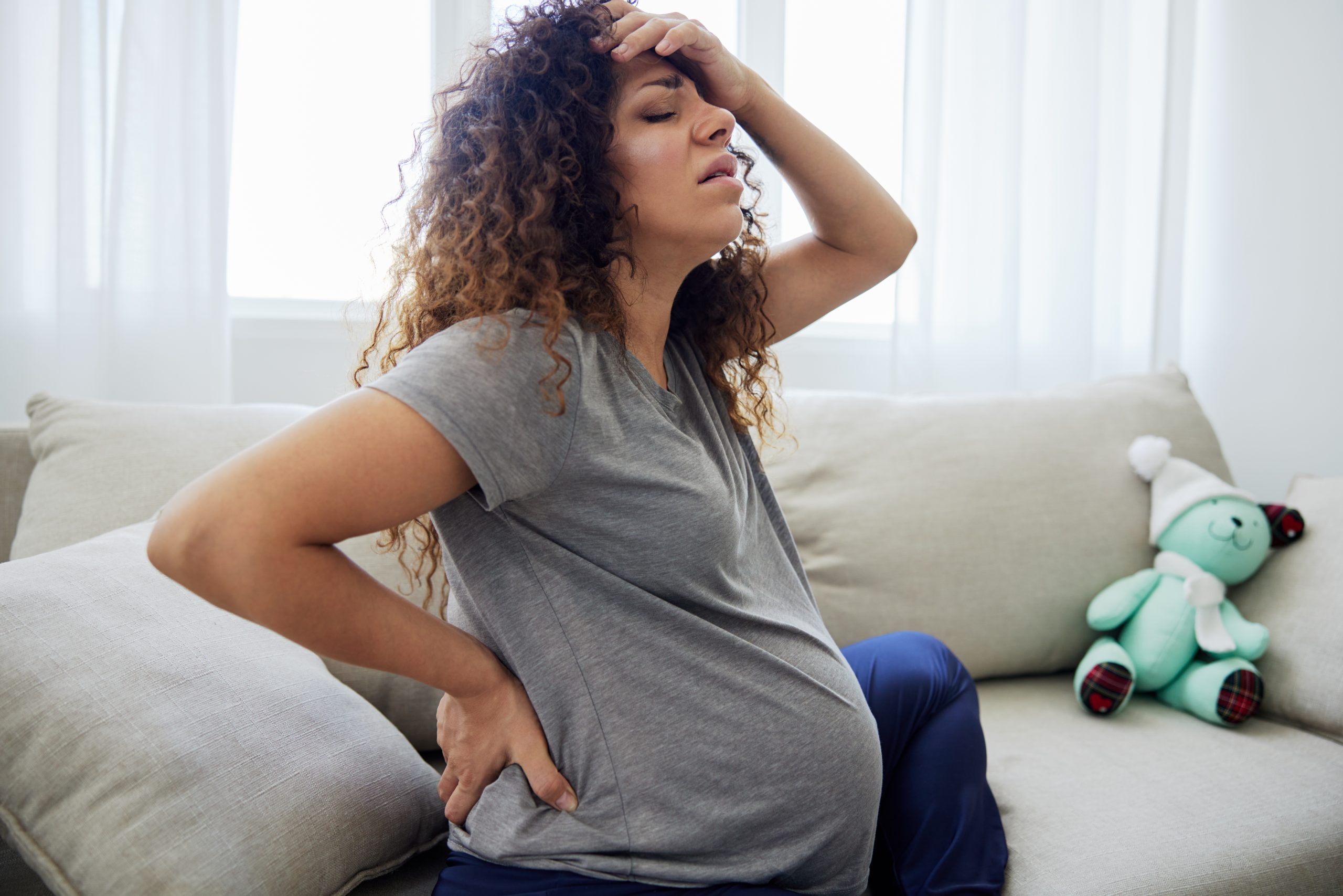 A pregnant woman with curly hair holds her head and back in discomfort, sitting on a beige sofa beside a green teddy bear. Bright white curtains frame the background.
