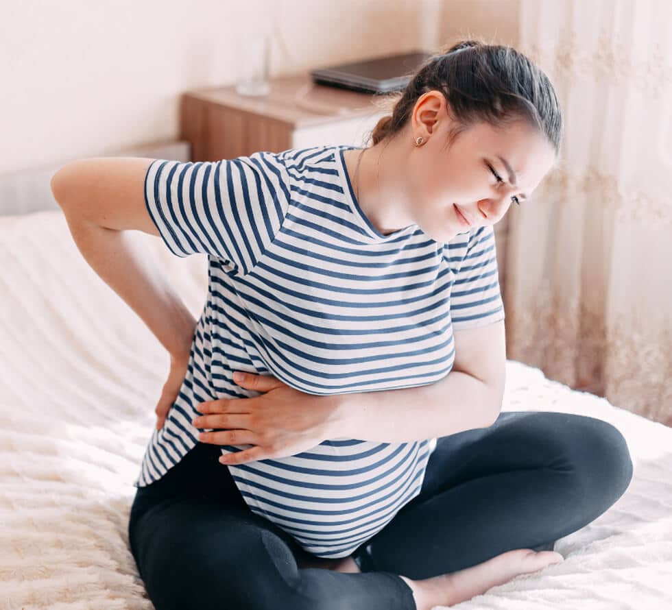 Pregnant woman sitting on a bed, holding her lower back and stomach, appearing uncomfortable. Room features a wooden nightstand with a closed laptop and sheer curtains.