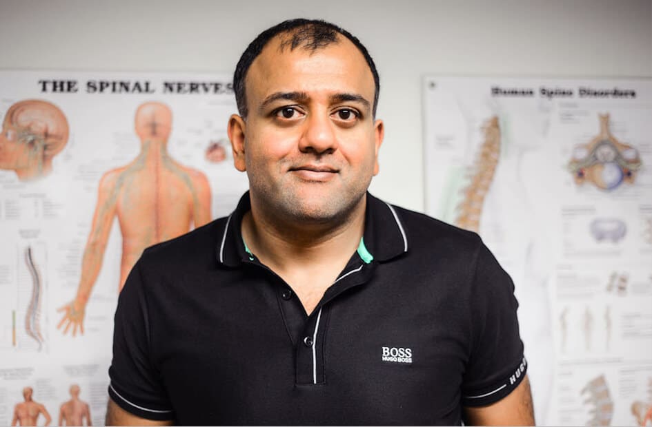 Avin stands in front of anatomical posters titled "THE SPINAL NERVES" and "Human Spine Disorders," wearing a black polo shirt with "BOSS" logo, in a clinical setting.