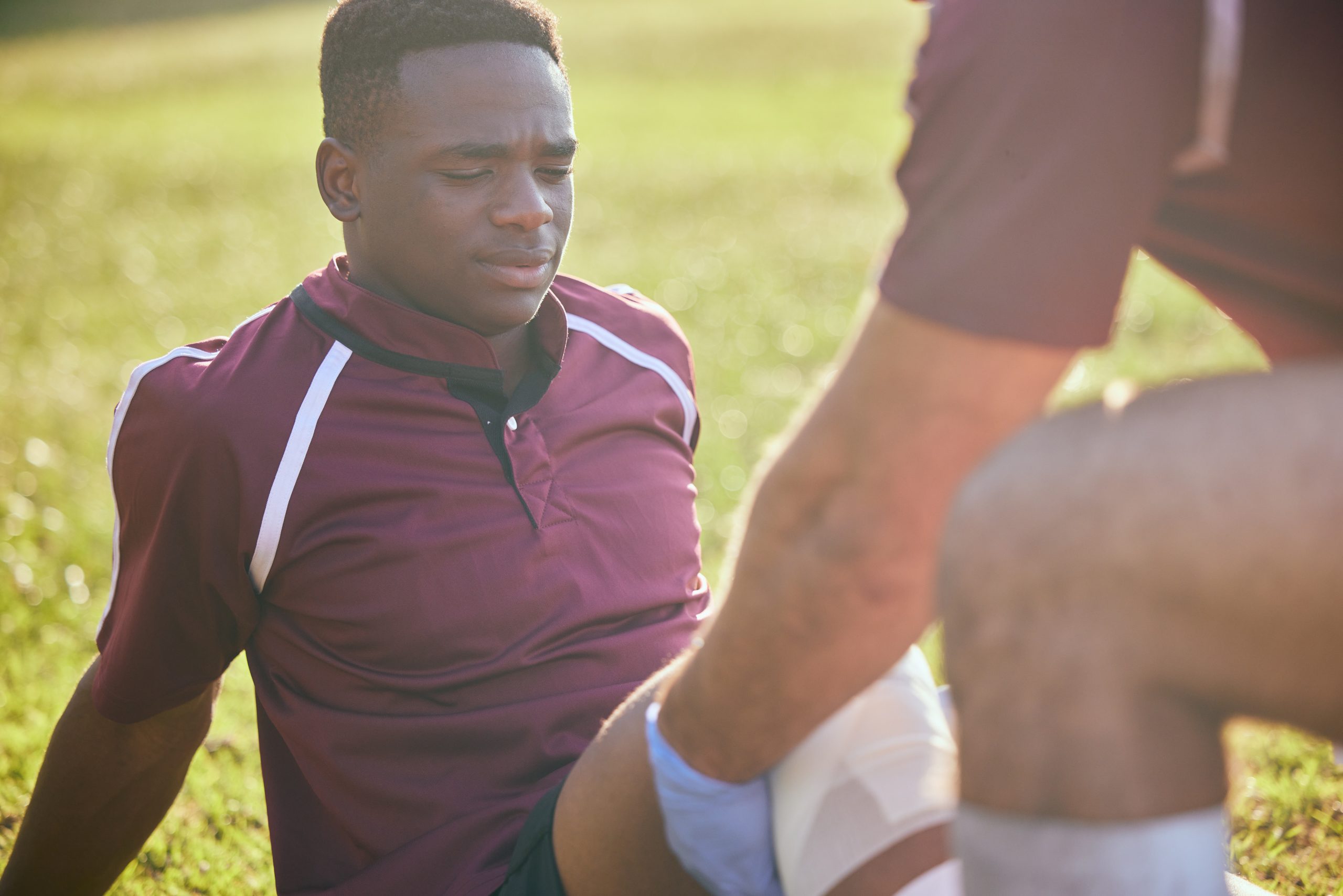 A seated person in a maroon sports jersey receives medical attention on their knee from another individual on a grassy field bathed in warm sunlight.