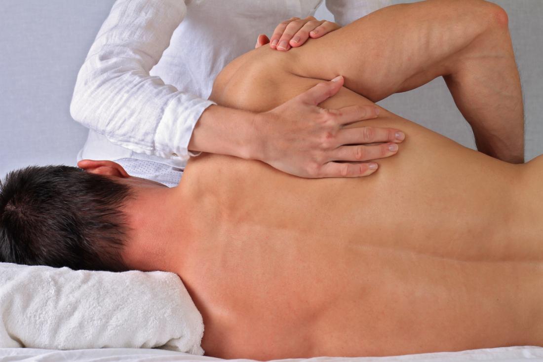 Person receiving a massage, lying face down on a white towel, while another person applies pressure to the back. The setting appears to be a spa or therapy room.