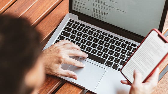 A person uses a laptop with both hands while holding a smartphone in one hand. They are seated at a wooden desk. The laptop screen displays text.