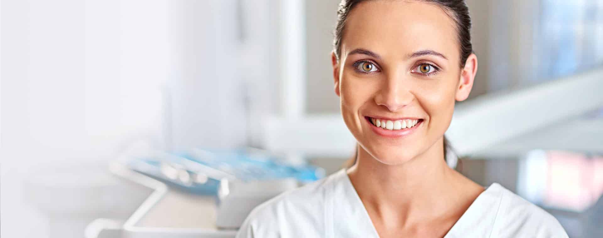 A person in a white uniform smiles warmly, situated in a well-lit, clinical setting with blurred background elements suggesting medical equipment.