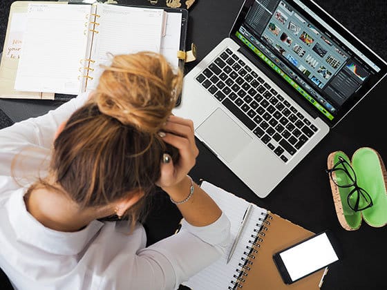 A person with tied-up hair holds their head while looking at a laptop displaying editing software. Nearby are open notebooks, a smartphone, and glasses on a dark desk.